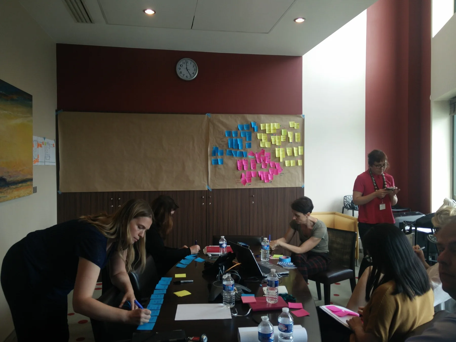 Workshop room at the bank: participants writing sticky notes around a table, kraft-paper research walls behind them